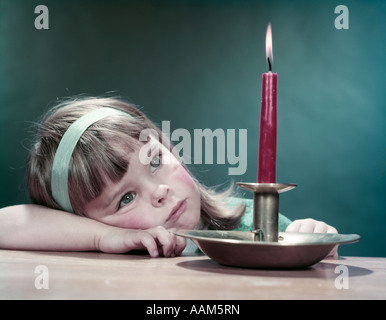 1950ER JAHRE MÄDCHEN WEHMÜTIG TRAURIG NACHDENKLICHE SCHLANKE KOPF AUF ARM AUF TABELLE BLICK AUF BRENNENDE ROTE KERZE IN MESSING HALTERUNG MAKE A WISH Stockfoto