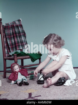 1950ER JAHREN LOCKIGES HAAR KLEINKIND SITZEN AM BODEN, DIE DURCH STUHL UND BABY PUPPE SPIELZEUG IHR PATENT LEDER SCHUHE ANZIEHEN Stockfoto