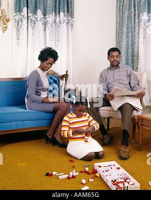 1960ER JAHRE AFRO-AMERIKANISCHE FAMILIE MUTTER VATER SITZEN LESUNG WOHNZIMMER MÄDCHEN TOCHTER SPIELEN STOCK SPIELZEUG LEGO RETRO Stockfoto