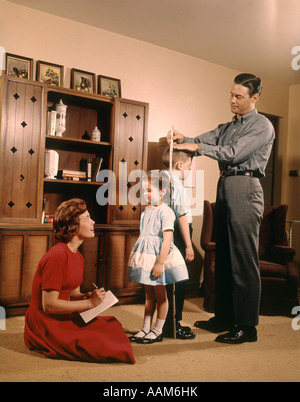 1950S 1960S VATER MUTTER SOHN TOCHTER MESSUNG HÖHE SCHWESTER & BRUDER FAMILIE INDOOR WOHNZIMMER AUFWACHSEN Stockfoto