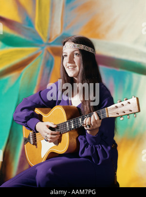 1970ER JAHRE JUNGE FRAU TRAGEN LILA KLEID UND PERLEN STIRNBAND SPIELT AKUSTISCHE GITARRE GEMUSTERTEN HINTERGRUND FOLK SONG Stockfoto