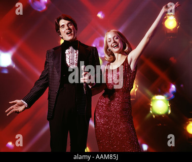 1970ER JAHREN UNTERHALTEN, FRAU UND MANN MIT MIKROFON SINGEN UND GESTIKULIEREN STROBOSKOPLICHT IM HINTERGRUND SMOKING KLEID Stockfoto