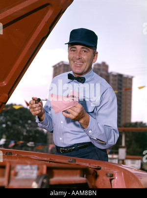 1960ER JAHRE MANN GAS SERVICE STATION MECHANISCHE ÜBERPRÜFUNG ÖL PEILSTAB UNTER AUTO KAPUZE LÄCHELND RETRO BLAUE UNIFORM HUT FLIEGE Stockfoto