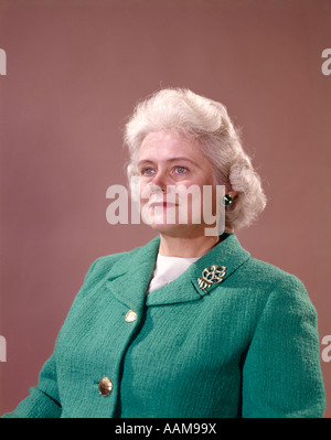 1960ER JAHRE PORTRAIT HALB LÄCHELND SENIOR WOMAN BEIGE HINTERGRUND GRAU SILBER HAAR TRAGEN GRÜNE STRICKJACKE GOLDKNÖPFEN BROSCHE Stockfoto