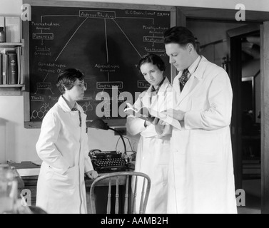 1930S 1940S BUCHEN SIE DREI WISSENSCHAFTLER IN WEIßEN LABORKITTELN BERATUNG EIN MANN ZWEI FRAUEN BLACKBOARD Stockfoto