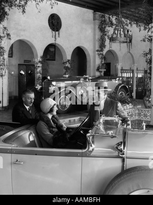 1920ER JAHREN FRAU IM FAHRERSITZ CABRIO MIT EHEMANN & VERKÄUFER STEHEN NEBEN MIETWAGEN IN AUSGEFALLENEN SHOWROOM Stockfoto