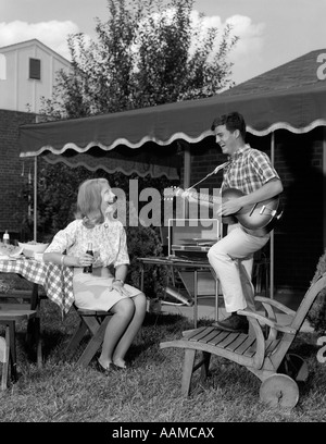 1960ER JAHRE TEENAGER-PAAR BOY SPIELEN GITARRE WOMAN SITTING ON GARTENMÖBEL MIT SOFTDRINK FLASCHE DRAUßEN IM HINTERHOF Stockfoto