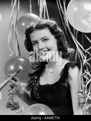 1940S 1950S LÄCHELNDE FRAU SCHWARZ PARTYKLEID PERLEN HALTEN SILVESTER PARTY NOISE MAKER LUFTSCHLANGEN LUFTBALLONS HINTERGRUND Stockfoto