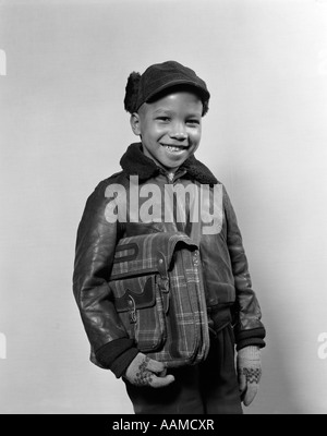 1940S 1950S AFRICAN AMERICAN BOY LÄCHELN AUF DEN LIPPEN TRAGEN WINTER JACKE HANDSCHUHE HOLDING BUCH SCHULTASCHE Stockfoto