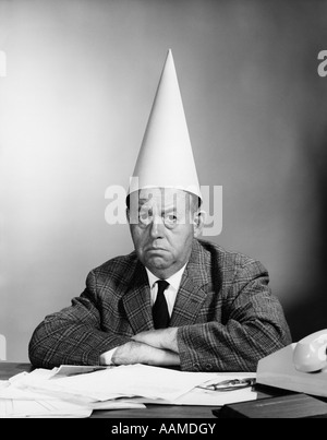 1960ER JAHREN UNGLÜCKLICH GESCHÄFTSMANN HINTER SCHREIBTISCH MIT DUNCE MÜTZE BLICK IN DIE KAMERA Stockfoto