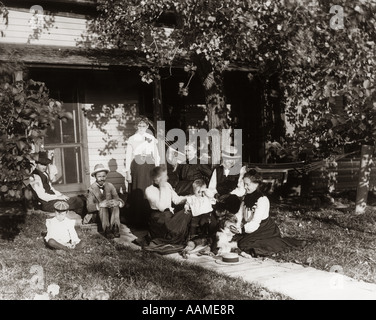 1890S 1900S WENDE DES JAHRHUNDERTS GROßFAMILIE SOMMER TAG SITZEN RASEN DES HAUSES MIT DREI GENERATIONEN & EIN HUND AUF VERANDA Stockfoto