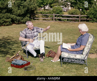 1960ER JAHRE ÄLTERES EHEPAAR SITZT IM HOF FRAU LESEN MENSCHEN ARBEITEN MIT ANGELGERÄT GEAR Stockfoto