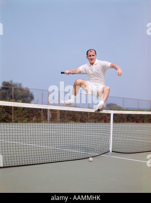 MITTE DER 1970ER-JAHRE ALTER MANN SPRINGT ÜBER NETZ AUF TENNISPLATZ GLATZENBILDUNG FITNESS AGILE WELLNESS LIFESTYLE Stockfoto