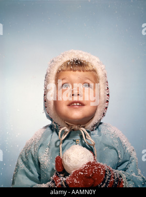 1960ER JAHRE MÄDCHEN HALTEN SCHNEE BALL BLICKTE ZU SKY HOFFENTLICH ROTE HANDSCHUHE WEIß STRICKEN HUT BLAU MANTEL Stockfoto