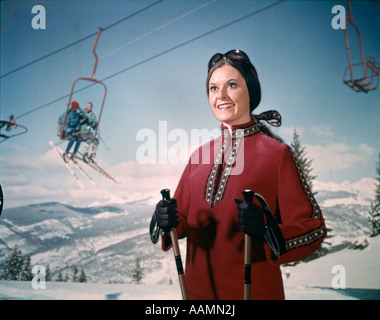 1970 1970S RETRO-FRAU IN WINTERKLEIDUNG UND SCHUTZBRILLEN HOLDING SKI POLEN SKI LIFT SCHNEEBERGE IM HINTERGRUND Stockfoto