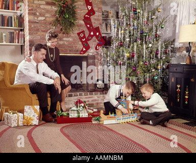 1960ER JAHRE FAMILIE IM WOHNZIMMER WEIHNACHTSBAUM JUNGS SPIELEN MIT SPIELZEUG MUTTER VATER SITZEN IM STUHL Stockfoto