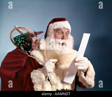 1950ER 1960ER JAHRE 1970ER JAHREN ÜBERRASCHT SANTA CLAUS MIT PLÜNDERUNG SPIELZEUG LESEN LISTE STUDIO INDOOR Stockfoto