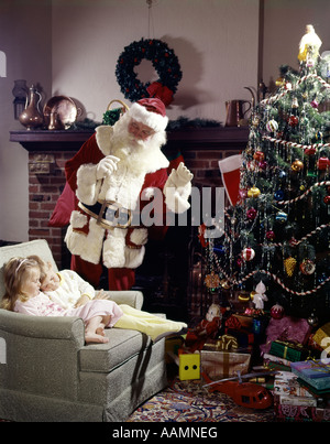 1960ER JAHRE SANTA CLAUS IM WOHNZIMMER IN DER NÄHE VON WEIHNACHTSBAUM WINKEN FÜR JUNGEN UND MÄDCHEN SCHLAFEN IM STUHL Stockfoto