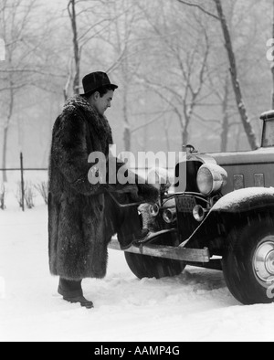 1930ER JAHRE MANN IN HUT UND WASCHBÄR PELZ, STEHENDE FUß AUF STOßSTANGE DES CHEVROLET ROADSTER INS STOCKEN GERATEN IN SCHNEESTURM Stockfoto