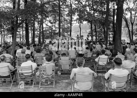 1950ER JAHRE MASSE GRUPPE MÄNNER FRAUEN SITZEN KLAPPSTÜHLE UNTER SCHATTEN SPENDENDEN BÄUMEN HÖREN SOMMER BAND KONZERT KLEINSTADT Stockfoto