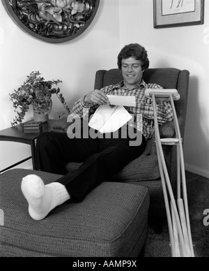 1970ER JAHRE JUNGER MANN IM SESSEL MIT EINEM BEIN AUFGESTÜTZT KRÜCKEN NAHEGELEGENEN BRIEFLESENDES INDOOR Stockfoto