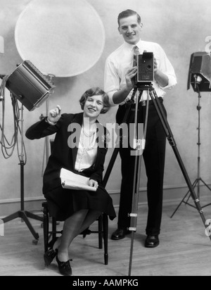 1920ER JAHREN FOTOGRAF HINTER DER KAMERA STEHT IM STUDIO MIT ASSISTENT SITZT NEBEN IHM HOLDING PAPIERKRAM Stockfoto