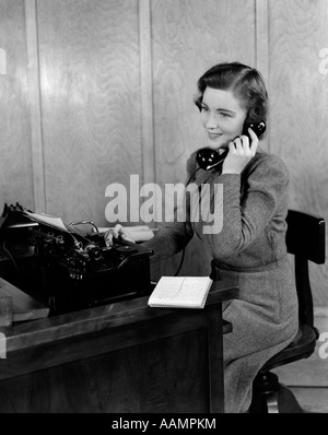 1940ER JAHRE LÄCHELNDE JUNGE FRAU SEKRETÄRIN ANGESTELLTE AN DER REZEPTION SITZEN AM SCHREIBTISCH, AM TELEFON SPRECHEN Stockfoto
