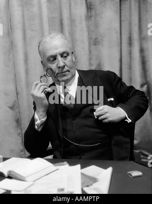 1930ER JAHRE MANN IN ANZUG & KRAWATTE SITZT AM SCHREIBTISCH HOLDING ZWICKER MIT NACHDENKLICHEN BLICK Stockfoto