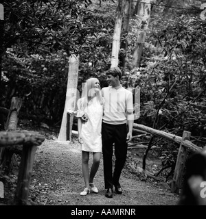 1960ER JAHRE JUNGES PAAR MANN FRAU BLOND ZU FUß AUßEN WEG BRÜCKE ROMANTIK DATE FLIRT WALD NATURPARK WALD Stockfoto