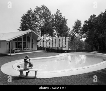 1960ER JAHRE BLOND IM BADEANZUG AUF STEINBANK SITZEND, AM ENDE DER NIERENFÖRMIGEN POOL Stockfoto