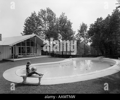 1950S 1960S BLONDE FRAU BADEANZUG SITZT AUF STEIN BANK BEACH-BALL IN DER NÄHE VON NIERE GEFORMTEN SWIMMINGPOOL Stockfoto