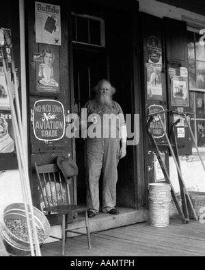 1930ER JAHRE ÄLTEREN BÄRTIGEN MANN IM OVERALL RAUCHT PFEIFE STEHT IM TOR DES GENERAL STORE Stockfoto