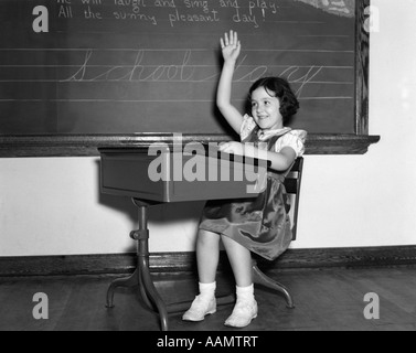 1930S 1940S LÄCHELNDE MÄDCHEN SITZEN AM SCHREIBTISCH HEBEN IHRE HAND Stockfoto