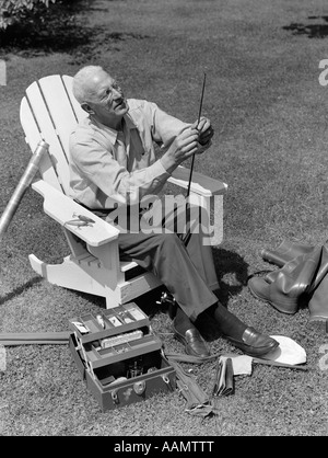 1950ER JAHRE ÄLTEREN MANN IM HINTERHOF SITZEN IM HOLZSTUHL EINFÄDELN LINIE DURCH ANGELRUTE MIT STIEFEL & TACKLE BOX ZU FÜßEN Stockfoto