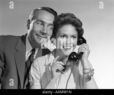 1950S 1960S FRAU AM TELEFON LÄCHELN MANN LEHNT SICH IN INNENRÄUMEN ZU HÖREN Stockfoto