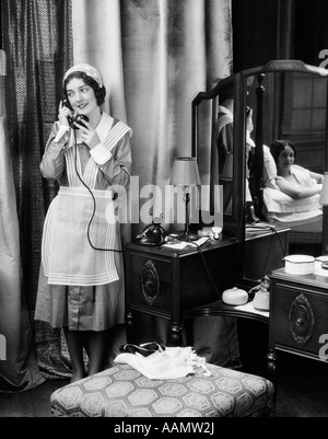 1920S 1930S MAID EINHEITLICHE GESPRÄCHE AM TELEFON VOR EITELKEIT TABELLE ANDERE FRAU PUTZT GILT ALS REFLEXION IM SPIEGEL Stockfoto
