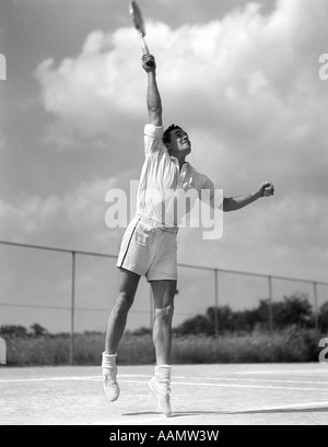 1930ER JAHRE MANN IN DER MITTE DIENEN SPIELEN TENNIS SCHWINGENDEN TENNISSCHLÄGER UM BALL SPRINGEN Stockfoto