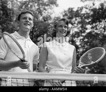 1960ER JAHREN LÄCHELND PORTRAIT PAAR HINTER NET HOLDING TENNISSCHLÄGER Stockfoto