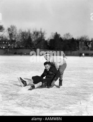 1920S 1930S ZWEI LÄCHELNDEN JUNGEN BLICK AUF KAMERA EISLAUFEN EIN JUNGE GEFALLEN ANDERE HOLTE IHN HERAUF Stockfoto