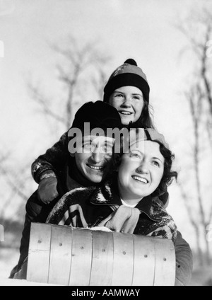 1930ER JAHREN SCHAL VATER MUTTER KIND AUF SCHLITTEN FACING KAMERA LÄCHELND HUT HAUBE Stockfoto