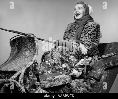 1950ER JAHRE LACHENDE FRAU IN LEOPARD HAUT PELZMANTEL FAHREN SCHLITTEN BELADEN MIT GESCHENKEN Stockfoto