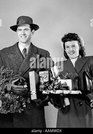 1940ER JAHREN PAAR LÄCHELND STEHEN IN WINTERJACKEN HALTEN VERPACKTE GESCHENKE UND EINEN KRANZ Stockfoto