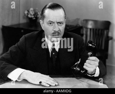 1930S 1940S PORTRAIT DER GESCHÄFTSMANN SITZT AM SCHREIBTISCH HOLDING BÜROTELEFON IN EINEN ERNSTEN GESICHTSAUSDRUCK SCHNURRBART Stockfoto