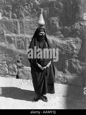 1920S 1930S ÄGYPTISCHE FRAU MIT KRUG AUF KOPF BALANCIERT TRAGEN TRADITIONELLE ARABISCHE MUSLIMISCHE GEWAND GESICHT VERSCHLEIERT BURKA Stockfoto