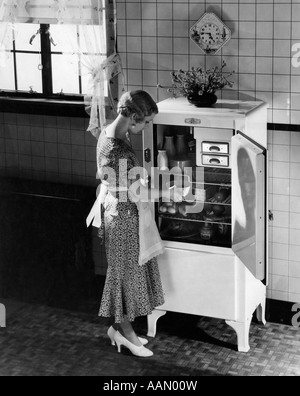 1920S 1930S BLONDE FRAU IN SCHÜRZE SEIDE PARTYKLEID HIGH HEELS EINNAHME GERICHT AUS ELEKTRISCHE KÜCHE KÜHLSCHRANK Stockfoto