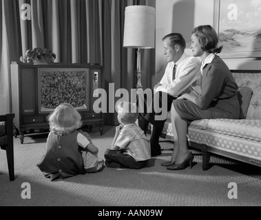 1960ER JAHRE FAMILIE SITZEN VIER IM WOHNZIMMER ANSCHAUEN FERNSEHEN MAMA PAPA AUF COUCH JUNGE MÄDCHEN AUF BODEN Stockfoto