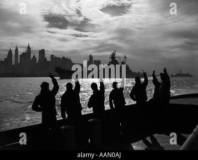 1940S 1950S SILHOUETTE SEGLER DECK DES SCHIFFES WINKEN GRUß AN VORBEI USN SCHLACHTSCHIFF IN DER NACHT VOR DER SKYLINE VON NEW YORK CITY Stockfoto