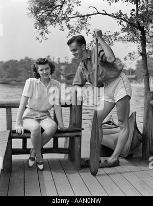 1940ER JAHRE MANN UND FRAU AM DOCK VON SEE FRAU SITZEN MANN STEHEND HOLDING RUDER KANU IM HINTERGRUND SOMMER FLIRT Stockfoto