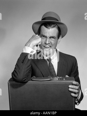 1960ER JAHREN ANGEWIDERT MANN MIT FINSTEREN BLICK AUF GESICHT TRAGEN HUT HEMD KRAWATTE ANZUG EINEN ELLBOGEN RUHEN AUF AKTENKOFFER BÖSE ÜBERRASCHUNG Stockfoto