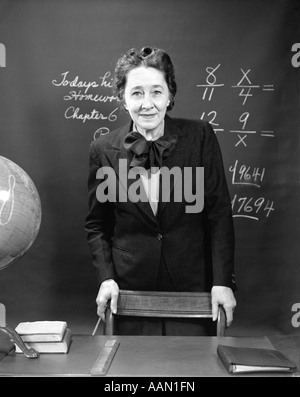 1940ER JAHREN LÄCHELND SCHULLEHRER STEHEN HINTER IHREM SCHREIBTISCH & VOR DEM SCHWARZEN BRETT MIT BRÜCHEN Stockfoto